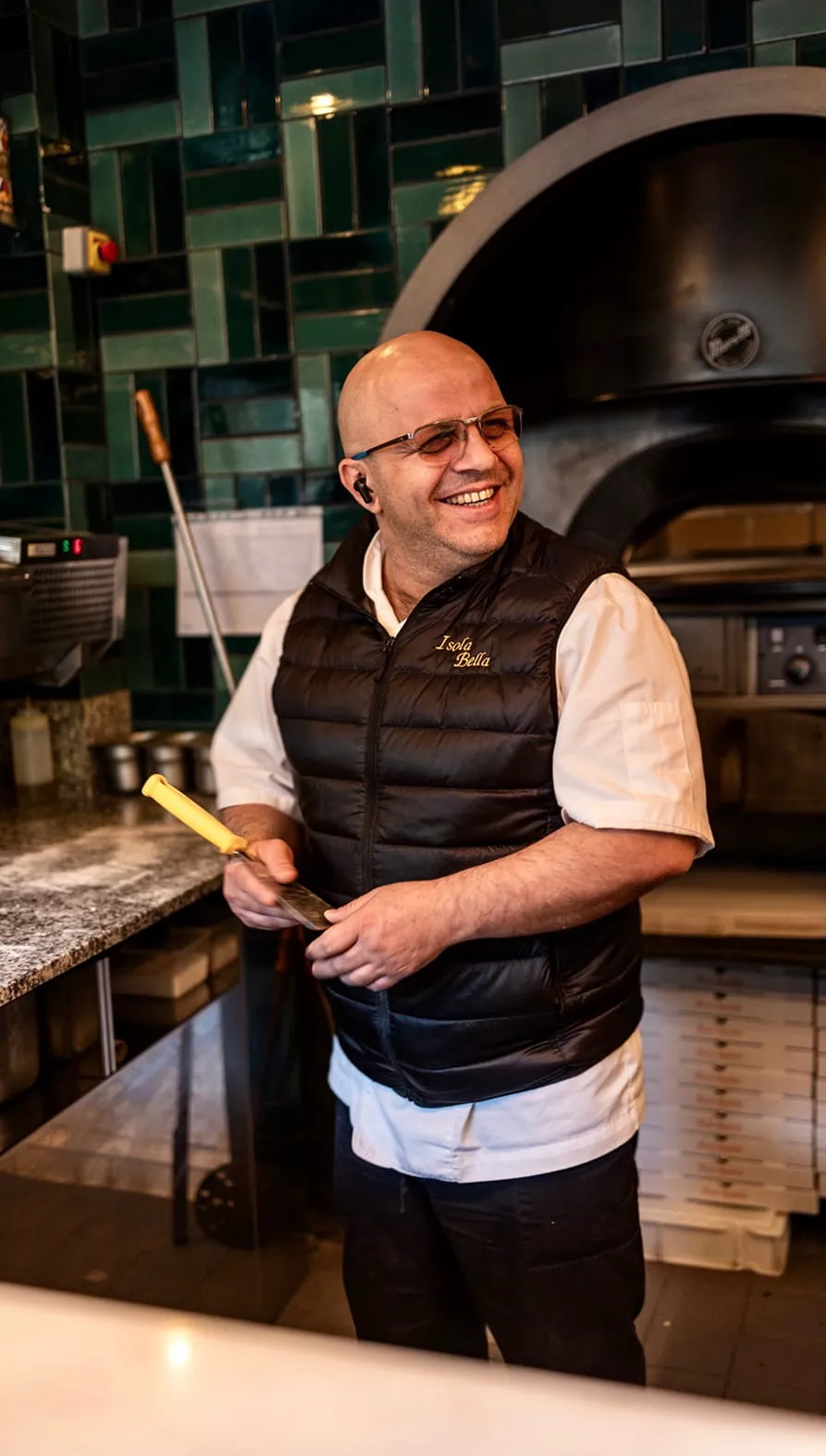 Un homme souriant portant une veste noire Isola Bella, debout dans une cuisine avec un four à pizza derrière lui.